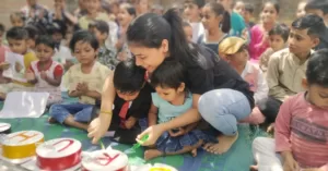 A young volunteer of aapke saath foundation helping two small children cut a cake while surrounded by a group of other children sitting on the ground, eagerly watching the birthday celebration organised by the NGO.