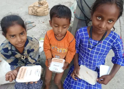 Two girls and one boy sitting on the road, holding food packets distributed by an AAPKE SAATH Foundation volunteer during the 'Food for Every Breath' campaign.
