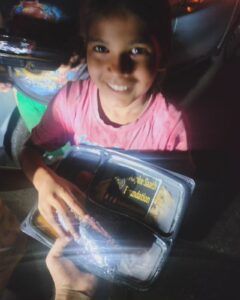 A young girl, around 7-9 years old, smiling as she receives a food plate from an AAPKE SAATH Foundation volunteer on a road at night.
