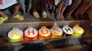 Children sitting on the stairs around six cakes spelling 'KHUSHI,' waiting to cut the cake.