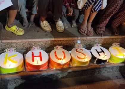 Children sitting on the stairs around six cakes spelling 'KHUSHI,' waiting to cut the cake.