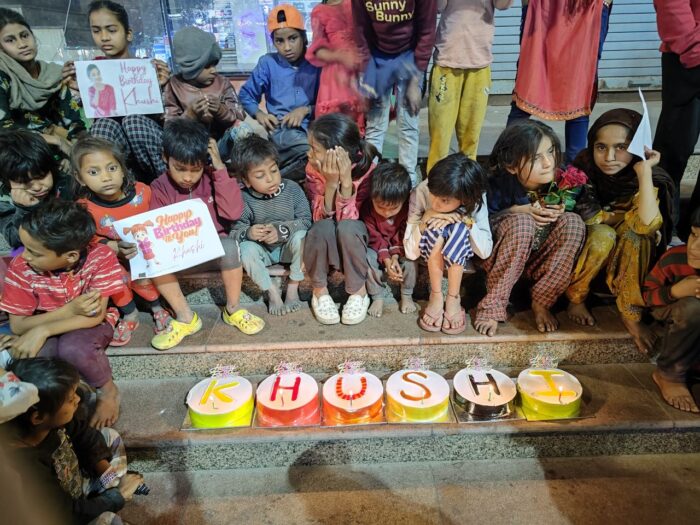 Children sitting on the stairs around six cakes spelling 'KHUSHI,' waiting to cut the cake. Some children hold a poster with a unique design that says 'Happy Birthday Khushi.