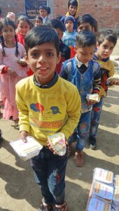 A donor distributing cake and food packets to children on their special day (birthday or anniversary). The children are lined up, smiling and holding cake and food packets in both hands. A banner in the background displays the NGO name 'AAPKE SAATH Foundation.